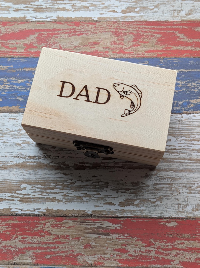 May include: A rectangular light-colored wooden box featuring a dark brown laser-engraved design on the lid. The engraving includes the word DAD in capital letters followed by a graphic illustration of a fish. The box has a metal latch closure.