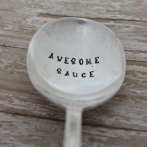 May include: A silver spoon with the words "AWESOME SAUCE" stamped on the bowl. The spoon has a simple, classic design and is set against a rustic, weathered wooden background.