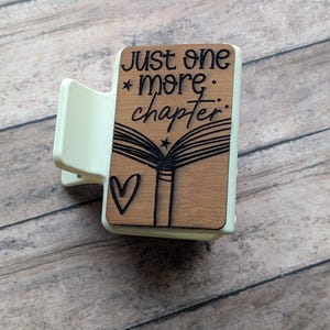 May include: A light green hair clip with a wooden accent featuring the text "just one more chapter" and a heart and book design. The clip is designed to hold hair in place.