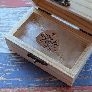 May include: A silver-toned metal fishing lure shaped like a guitar pick, engraved with the text I'LL ALWAYS BE YOUR FISHING BUDDY, featuring a treble hook and swivel, presented inside a small, light-colored wooden box with a metal latch.