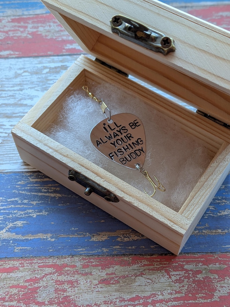 May include: A silver-toned metal fishing lure shaped like a guitar pick, engraved with the text I'LL ALWAYS BE YOUR FISHING BUDDY, featuring a treble hook and swivel, presented inside a small, light-colored wooden box with a metal latch.