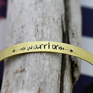 May include: A gold-toned metal bracelet with the word "warrior" engraved on it, along with an arrow and leaves on either side.