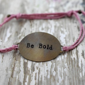 May include: A pink braided cord bracelet with a brass oval charm that says "Be Bold".
