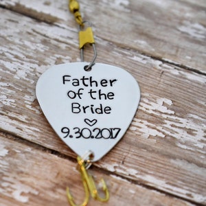 May include: A silver-coloured metal guitar pick shaped fishing lure engraved with the text Father of the Bride, a small heart symbol, and the date 9.30.2017. The lure features a gold-toned treble hook at the bottom and a gold swivel connector at the top.