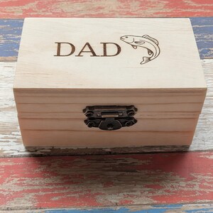 May include: A rectangular light-colored wooden box with a hinged lid featuring the laser-engraved text DAD and a graphic of a fish. The box is secured with a dark bronze-colored metal latch centered on the front panel of the wooden container.