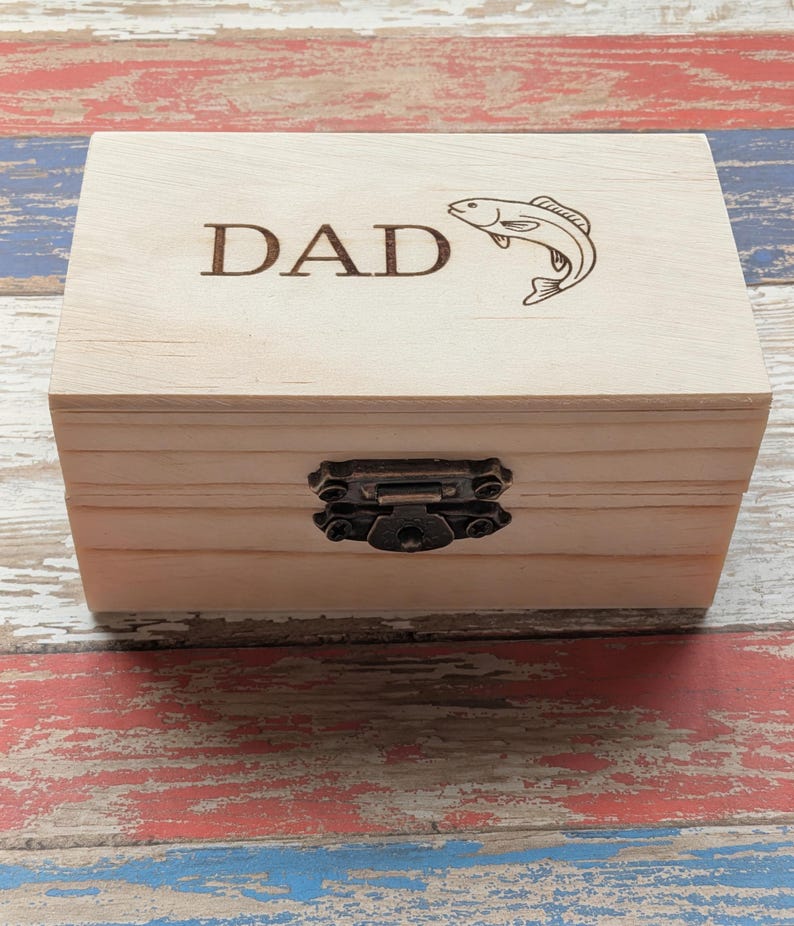May include: A rectangular light-colored wooden box with a hinged lid featuring the laser-engraved text DAD and a graphic of a fish. The box is secured with a dark bronze-colored metal latch centered on the front panel of the wooden container.