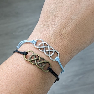 May include: Two infinity symbol bracelets on a wrist. One bracelet is black with a gold infinity symbol and the other is light blue with a silver infinity symbol.