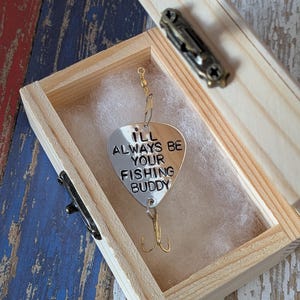 May include: A silver-colored metal fishing lure shaped like a guitar pick, engraved with the text "I'LL ALWAYS BE YOUR FISHING BUDDY." The lure features a gold-toned treble hook at the bottom and a swivel connector at the top, resting inside a small wooden box.