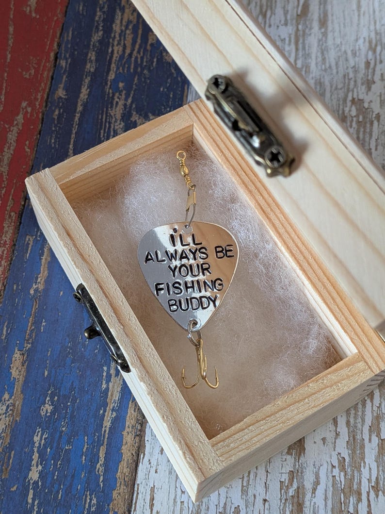 May include: A silver-colored metal fishing lure shaped like a guitar pick, engraved with the text "I'LL ALWAYS BE YOUR FISHING BUDDY." The lure features a gold-toned treble hook at the bottom and a swivel connector at the top, resting inside a small wooden box.