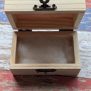 May include: A small, rectangular, light-colored wooden box with an open lid. The box features a dark bronze-toned metal latch mechanism on the front and a soft, white, fibrous padding lining the interior base of the container.