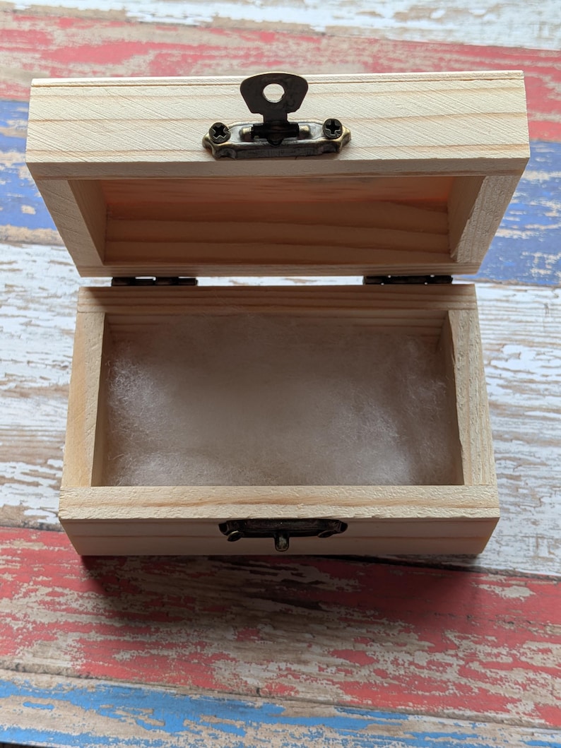 May include: A small, rectangular, light-colored wooden box with an open lid. The box features a dark bronze-toned metal latch mechanism on the front and a soft, white, fibrous padding lining the interior base of the container.