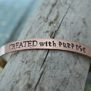 May include: A copper-coloured bracelet with the words "CREATED with PURPOSE" engraved on it.