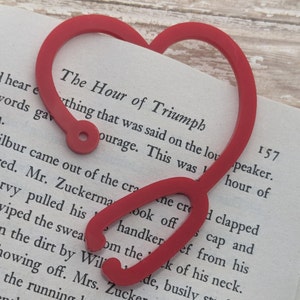 May include: A red heart-shaped bookmark with a small hole at the top. The bookmark is on top of a book with the text "The Hour of Triumph" visible.