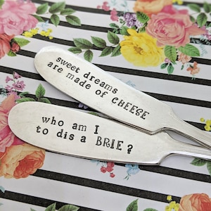 May include: Two silver cheese spreaders with engraved sayings. The top spreader reads "Sweet dreams are made of cheese". The bottom spreader reads "Who am I to dis a brie?"