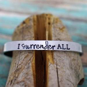 May include: A silver metal bracelet with the words "I surrender ALL" engraved on it. The bracelet is resting on a piece of weathered wood.