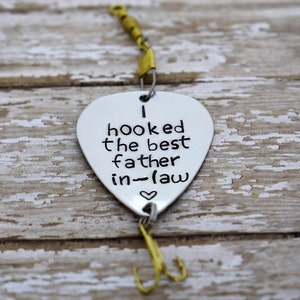May include: A silver-coloured metal fishing lure shaped like a guitar pick, featuring the stamped black text "I hooked the best father-in-law" with a small heart symbol. The lure includes a brass-coloured swivel at the top and a gold-toned treble hook.