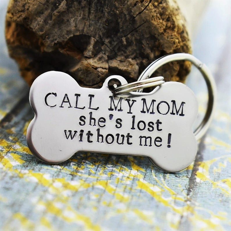 Op de afbeelding: Een zilverkleurige hondenpenning in de vorm van een bot met de gegraveerde tekst "CALL MY MOM she's lost without me!". De penning is bevestigd aan een zilveren splitring.