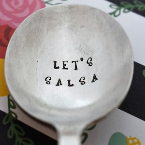May include: A silver spoon with the words "Let's Salsa" stamped on the bowl. The spoon is on a black and white striped background with floral designs.