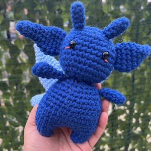 May include: A blue crocheted plush toy with large eyes and a small tail. The toy is being held in a person's hand.