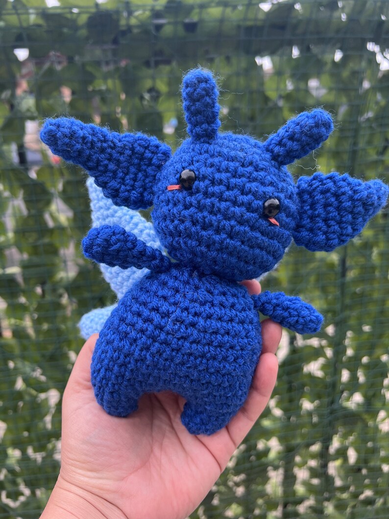 May include: A blue crocheted plush toy with large eyes and a small tail. The toy is being held in a person's hand.