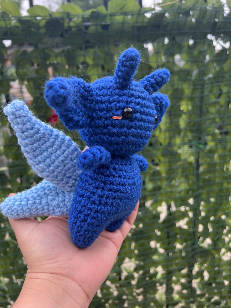 May include: A blue and light blue crocheted stuffed animal with a wing. The animal has a black eye and a pink blush.