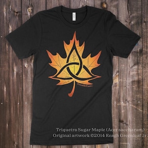 May include: Black t-shirt featuring a stylized orange and yellow maple leaf design with a triquetra knot in the center. The text "Triquetra Sugar Maple" and artist's name are visible. The shirt is made of a soft material.