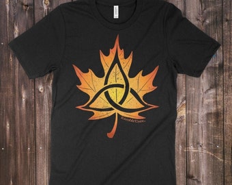 Könnte beinhalten: Schwarzes T-Shirt mit einem detaillierten orange-gelben Ahornblatt-Design, das einen Triquetra-Knoten enthält. Der Text "Triquetra Sugar Maple" und der Name des Künstlers sind sichtbar. Das T-Shirt ist aus weichem Material.