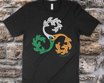 May include: Black t-shirt with a green, white, and orange triquetra design featuring oak leaves. The design is labeled "RambleTree (Quercus alba)" and "Original artwork ©2016 Reagh Greenical Art".
