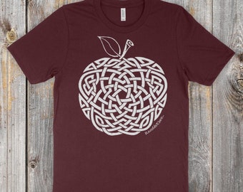 Op de afbeelding: Een maroon t-shirt met een wit Keltisch knoopontwerp van een appel. De tekst "Raddle Creek" staat onderaan het ontwerp. De tekst "Morgan's Apple (Malus domestica)" en "Original artwork ©2018 Reagh Greenleaf Jr." staat onderaan het shirt.