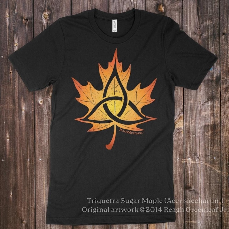 May include: Black t-shirt featuring a stylized orange and yellow maple leaf design with a triquetra knot in the center. The text "Triquetra Sugar Maple" and artist's name are visible. The shirt is made of a soft material.