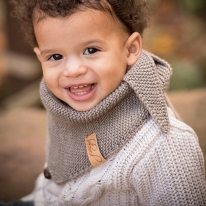 May include: A young child with curly brown hair wearing a white knitted sweater with a brown knitted scarf. The scarf has a brown leather tag with the text "Baby Brand" on it.