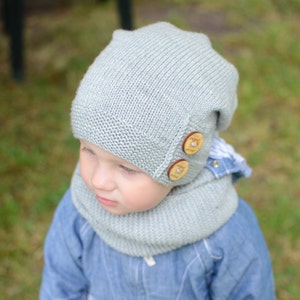 May include: A gray knitted hat with wooden buttons and a matching gray knitted scarf. The hat has a slouchy fit and the scarf is a cowl style.