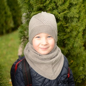 May include: A young person wearing a gray knitted hat and scarf. The hat has a unique design with a pointed top and a flap that covers the ears. The scarf is a long, flowing style that wraps around the neck.