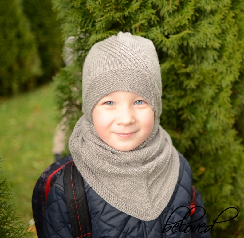 May include: A young person wearing a gray knitted hat and scarf. The hat has a unique design with a pointed top and a flap that covers the ears. The scarf is a long, flowing style that wraps around the neck.