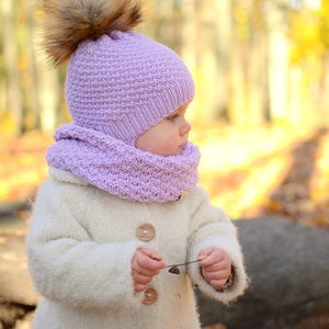 K&ouml;nnte beinhalten: Ein kleines Kind tr&auml;gt einen wei&szlig;en Strickmantel mit Holzkn&ouml;pfen, eine lilafarbene Strickm&uuml;tze mit einem braunen Pelzbommel und einen passenden lilafarbenen Strickschal. Das Kind steht in einer Waldlandschaft mit heruntergefallenen Bl&auml;ttern auf dem Boden.
