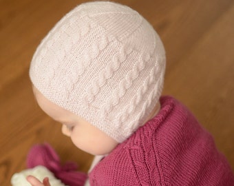 May include: A baby wearing a pink knitted hat with a cable knit pattern and a burgundy knitted sweater.