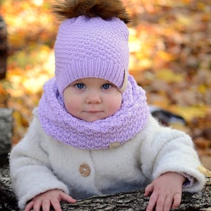 K&ouml;nnte beinhalten: Ein Baby tr&auml;gt eine lilafarbene Strickm&uuml;tze mit einem braunen Bommel und einen passenden lilafarbenen Strickschal. Das Baby sitzt auf einem Baumstamm mit einem verschwommenen Hintergrund aus Herbstlaub.