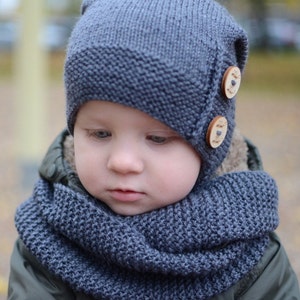 Könnte beinhalten: Ein Baby trägt eine blaue Strickmütze und einen passenden Schal. Die Mütze hat zwei Holzknöpfe mit Text darauf.