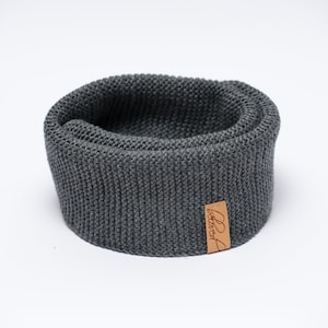 May include: A dark gray knitted neck warmer with a rolled edge. A small, rectangular brown leather tag with the word "beloved" is attached to the front. The neck warmer is made of a thick, textured yarn.