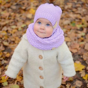 K&ouml;nnte beinhalten: Ein kleines Kind tr&auml;gt einen wei&szlig;en Strickmantel, eine lavendelfarbene Strickm&uuml;tze mit einem braunen Pelzbommel und einen lavendelfarbenen Strickschal. Das Kind steht in einem Haufen Herbstlaub.