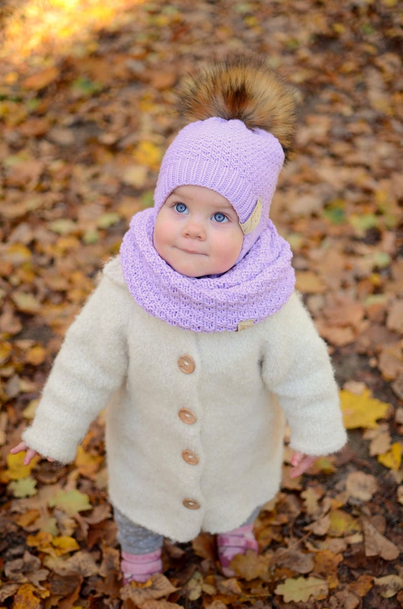 K&ouml;nnte beinhalten: Ein kleines Kind tr&auml;gt einen wei&szlig;en Strickmantel, eine lavendelfarbene Strickm&uuml;tze mit einem braunen Pelzbommel und einen lavendelfarbenen Strickschal. Das Kind steht in einem Haufen Herbstlaub.
