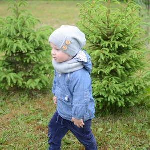 May include: A young child wearing a light blue jacket, dark blue pants, a gray knitted hat with wooden buttons, and a gray scarf. The child is standing in a grassy area with green trees in the background.