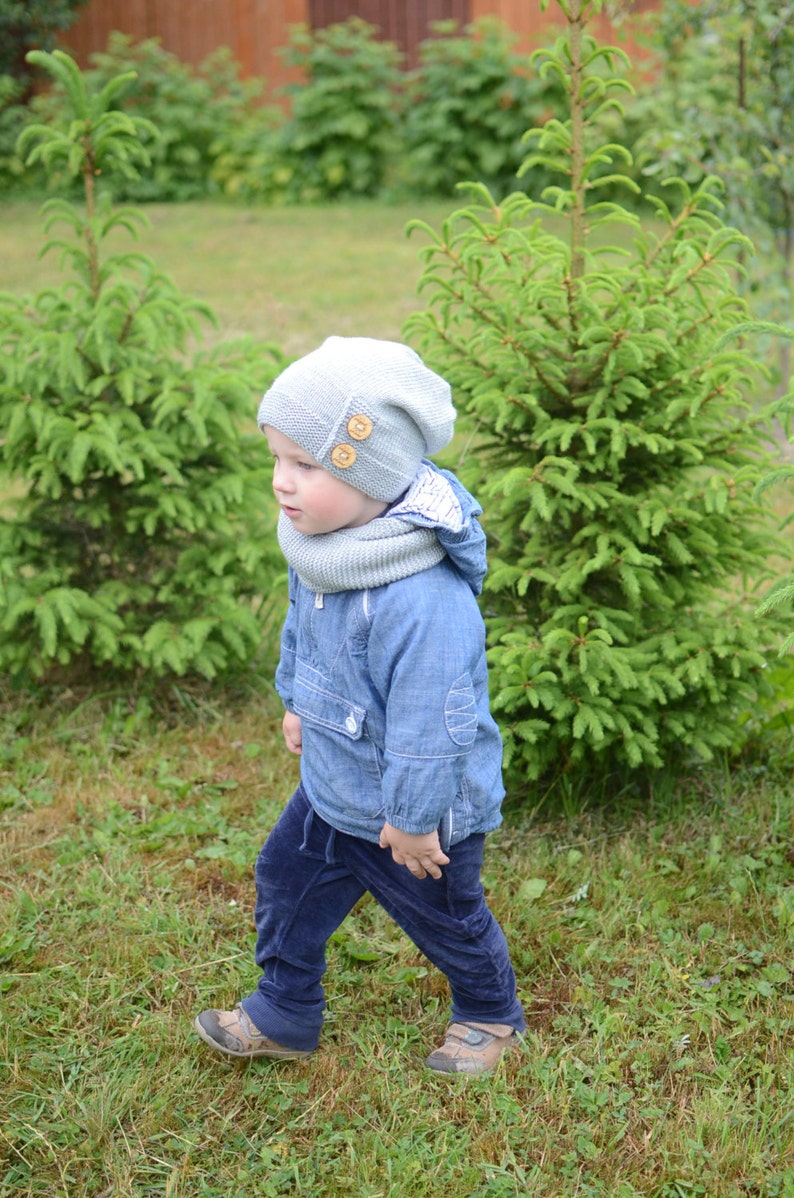 May include: A young child wearing a light blue jacket, dark blue pants, a gray knitted hat with wooden buttons, and a gray scarf. The child is standing in a grassy area with green trees in the background.