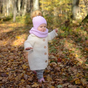 K&ouml;nnte beinhalten: Ein kleines Kind tr&auml;gt einen wei&szlig;en Mantel, eine lilafarbene Strickm&uuml;tze mit einem braunen Bommel und einen lilafarbenen Strickschal und geht durch einen Wald mit B&auml;umen und heruntergefallenen Bl&auml;ttern.