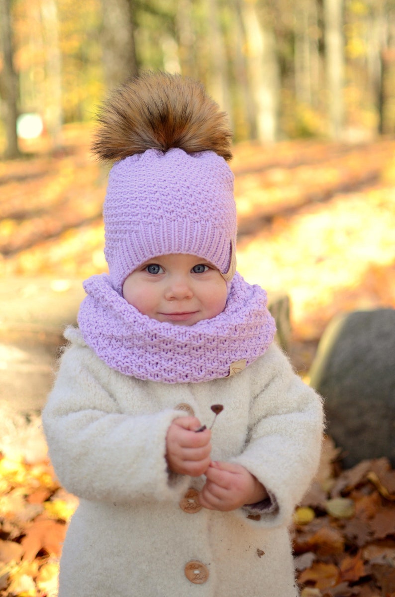 K&ouml;nnte beinhalten: Ein kleines Kind tr&auml;gt einen wei&szlig;en Strickmantel mit einer lilafarbenen Strickm&uuml;tze und einem Schal. Die M&uuml;tze hat einen braunen Pelzbommel oben. Das Kind h&auml;lt einen kleinen Zweig in der Hand.