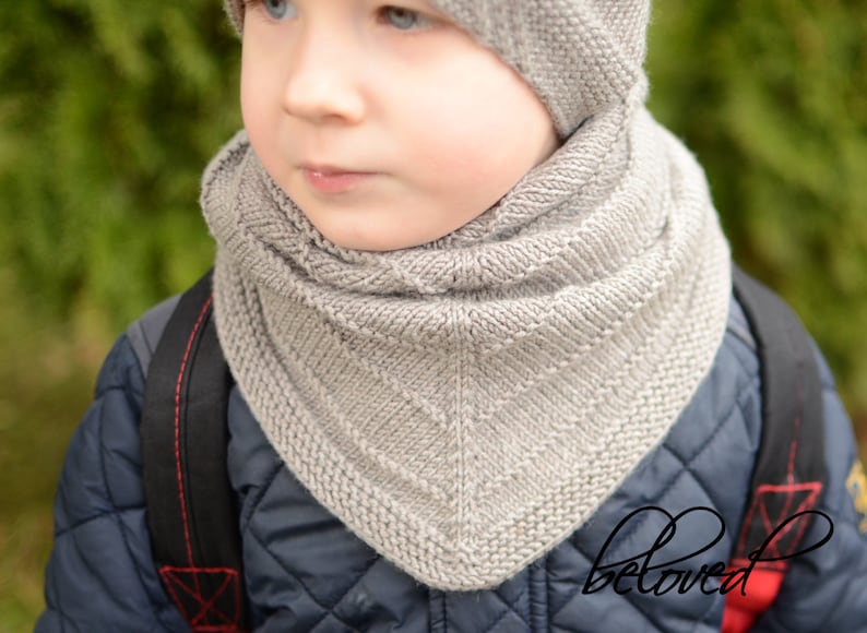 May include: A close-up of a child wearing a gray knitted scarf and hat. The scarf is folded over and has a diamond pattern. The hat is pulled down over the child's ears.
