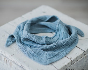 Könnte beinhalten: Ein hellblauer gestrickter Schal mit einem strukturierten Muster. Der Schal ist gefaltet und auf einer weißen Holzoberfläche drapiert.