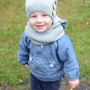 May include: A young child wearing a gray knitted hat with buttons and a matching scarf. The child is wearing a blue denim jacket and blue jeans. The child is standing on a path with green grass in the background.