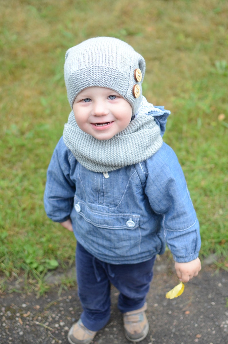 May include: A young child wearing a gray knitted hat with buttons and a matching scarf. The child is wearing a blue denim jacket and blue jeans. The child is standing on a path with green grass in the background.