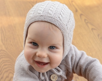 May include: A baby wearing a knitted beige hat with a cable knit pattern and a matching knitted cardigan. The baby is smiling and looking up at the camera.